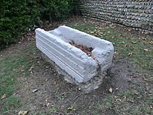 Sarcophage pris en contre-plongée, en pierre blanche, posé sur la terre. La paroi en largeur est endommagée, celle en longueur est rainurée.