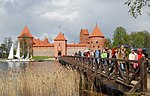 Red brick castle in the background. Lake in front. A wooden bridge with tourists. Sailboats on the lake.