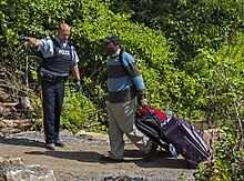 A bald uniformed police officer wearing black gloves and a blue flack jacket with "POLICE" written on it in white letters at left points in that direction while a man on the right in a blue and brown striped sweater wearing a baseball cap pulls a wheeled suitcase behind him on a dirt pathway with shrubs behind him