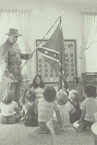 Black and white photo of man in confederate army uniform holding confederate flag in a classroom with children seated around him