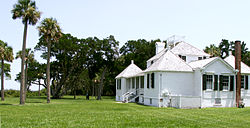 Color photo of the owner's house from a diagonal view, showing a large white structure with black shutters, two gabled pavilions separated by a porch, a taller larger gable in the center, a small gable roof to the right of the closest pavilion, and a partially obscured pavilion with a gabled roof and chimney. A lawn and bent oak and palm trees surround the house