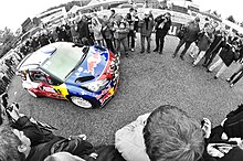 Montage photographique avec déformation sphérique et décor en noir et blanc, montrant la Citroën DS3 WRC de Sébastien Loeb entre deux rangés de spectateurs.