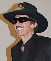 Richard Petty sporting a black cowboy hat and black sunglasses and a black suit inside a building