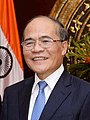 a smiling nearly bald man, wearing glasses, a suit and a red tie, back him is flag of India