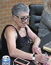 Shirley Muldowney wearing eye glasses and signing autographs for fans on a black table