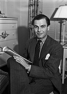 A Caucasian man wearing a suit, sitting on a chair with legs crossed, facing slightly left, and holding a newspaper in his lap