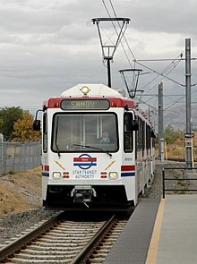 A white and red train with the driver visible approaches a station. The overhead wires powering the train are visible with overcast skies and mountains providing the backdrop.