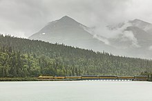 Paysage de montagnes et de forêts dans de la grisaille. Un train rayé d'une ligne jaune franchit un viaduc sur un lac et longe ce dernier.