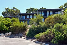 Three dark grey cube-like buildings, each with 2 storeys, are surrounded by native plants.