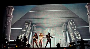 A female blond performer standing with two dancers in front of two giant LED screens, which has an image of a pyramid.