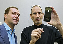 Shoulder-high portrait of two middle aged men, the one on left wearing a blue dress shirt and suitcoat, the one on right wearing a black turtleneck shirt and with his glasses pushed back onto his head and holding a phone facing them with an Apple logo visible on its back