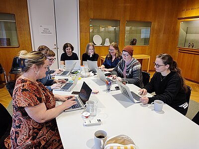 Sju personer runt ett konferensbord, med varsin dator framför sig.