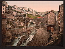 Carte postale ancienne colorisée montrant en contre-plongée la partie nord de la vallée en période de basses eaux, au centre un pont roman à 2 arches traverse la rivière, pont et maisons sont construits en pierres de schistes brunes.