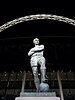 Statue of a footballer with arms crossed and his left leg resting on a football, with Bobby Moore written on the plinth, beneath the arch of the new Wembley stadium