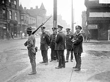 people standing on the street, one is armed with a rifle