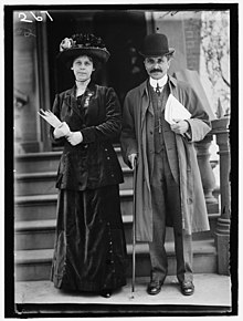 A white woman and a Persian man standing next to each other in front of stairs. Both are holding papers. She wearing a large brimmed hat; he is wearing a bowler hat.