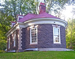 A stone building with red metal roof and a rounded projection in front. It is sheltered by trees to the rear.