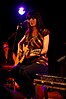 A woman with long black hair playing a guitar and singing in front of a microphone