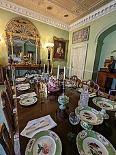 An 18th century dining room with a long table holding an array of ceramics, silverware and glassware.