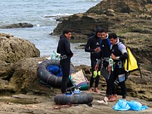 A croup of three divers dressed in wetsuits standing on a rocky shore with the sea in the background. On the ground are inflated truck inner tube floats with nets to support their catch