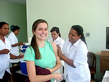 Uśmiechnięta kobieta w zielonej bluzce otrzymuje szczepionkę przeciwko różyczce. W tle krzątają się trzy inne kobiety.
