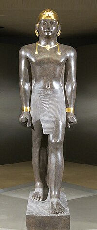 Statue of Taharqa. His name appears on the center of his belt: 𓇿𓉔𓃭𓈎 (tꜣ-h-rw-q, "Taharqa"). The statue is 2.7 meters tall. Taharqa has a striding pose, the arms held tight, and holds the mekes staff. He wears a pleated kilt called shendjyt and on the head the double-uraeus skullcap, possibly signifying the double rule over Nubia and Egypt.[1] (Louvre Museum, color reconstruction of the jewelry through pigment analysis).[2]