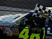 Jimmie Johnson holding a checkered flag in his left hand outside a left-hand side race car window