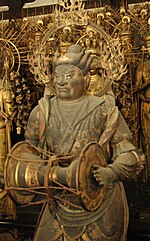 Un des vingt-huit assistants. Vue de trois-quarts d'une statue portant un tambour à main en face. Une auréole est visible autour de sa tête.