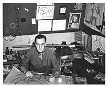 Stan Lee sits in a office, with several drawings on the background. He is sitting down in front of a table; on that table he is drawing an image.