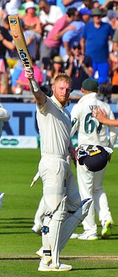 Un homme en tenue de joueur de cricket, lève sa batte vers le ciel. Il tient son casque dans l’autre main.