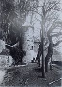 Vue ancienne de la tour d'un château contre une paroi rocheuse, avec une cour plantée de quelques arbres à l'avant.