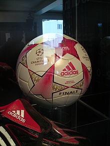 Sepak bola di kabinet perspex. Bola didominasi putih, bermotif bintang merah dengan detail emas. Di latar depan adalah sepatu sepak bola hitam dengan trim merah dan putih.