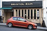 Photo d'un bâtiment avec de longues fenêtres étroites et un auvent au-dessus de l'entrée. Un panneau indique « BRISTOL RAM ». Une voiture rouge est stationnée à l'avant sur une rue en pente vers la droite.