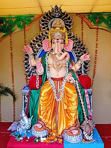 Attired in an orange dhoti, an elephant-headed man sits on a large lotus. His body is red in colour and he wears various golden necklaces and bracelets and a snake around his neck. On the three points of his crown, budding lotuses have been fixed. He holds in his two right hands the rosary (lower hand) and a cup filled with three modakas (round yellow sweets), a fourth modaka held by the curving trunk is just about to be tasted. In his two left hands, he holds a lotus in the upper hand and an axe in the lower one, with its handle leaning against his shoulder.