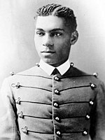 Cadet Henry O. Flipper in his West Point cadet uniform. It has three large round brass buttons left, middle and right showing five rows. The buttons are interconnected left to right and vice versa by decorative thread. He is wearing a starched white collar and no tie. He is a lighter colored African-American with plated corn rows of neatly done hair. He is facing the camera and looking to the left of the viewer.