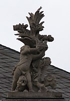 54. Trophäe – Baumstamm, Löwe, Widder, Putto als Kronenhalter.