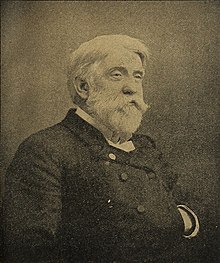 A white-haired man with a beard and mustache, facing left. He is wearing a white shirt, black vest, and black jacket