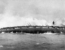 Una grande nave da guerra si sta rovesciando sul fianco, i marinai affollano il bordo emerso della nave, aggrappandosi disperatamente alle reti antisiluro. Sullo sfondo una enorme nube di vapore si leva dal relitto.