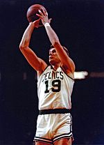 An older man, wearing a black suit, is sitting on the side of a basketball court.