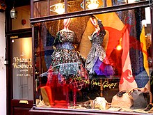 Frontale Farbfotografie von einem beleuchteten Schaufenster mit dunkler Holzumrandung. Zwei Schaufensterpuppen mit extravaganten Kleidern sind von Accessoires und einer glitzernden Leinwand umgeben. Auf dem Schaufenster ist die rote Silhouette von einem Teufel mit der goldenen Schrift „Wake up, Cave Girl“ abgebildet.