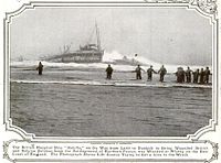 Black and white photo of a ship sinking into the water, with multiple people running towards it.