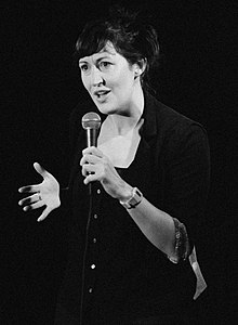 Black-and-white photo of Celia Pacquola standing onstage, speaking into a microphone