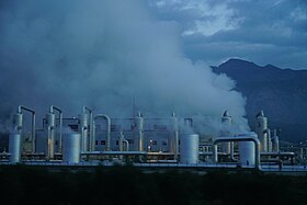 Lots of steam rises above metal pipes and vertical cylinders, with a low building and mountains in the background.