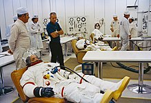 1971年7月26日，阿波罗15号发射前，几位身穿太空服的人躺在沙发上，其他人站在一旁。德克·斯莱顿（蓝色衣服）看着斯科特（离镜头最近的人）在穿好衣服后休息