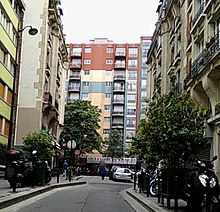 Photo couleur de la façade d'un immeuble de dix étages (fond de l'image), vue d'une rue (premier plan) bordée par des bâtiments d'habitations de quatre à cinq étages.