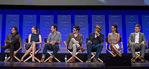 Seven people onstage in directors' chairs, looking right