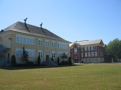 A yellow building made of wooden planks with a black tar roof, adjacent to a larger red brick building also with a black tar roof. The foreground is a trimmed grass field with a large tree on the side; the background is a clear blue sky.