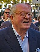 An elderly white man with receding white hair is wearing glasses. He is outdoors with various people in the background, suggesting a crowd. He is smiling, his mouth open. He is wearing a shirt with a blue jacket.