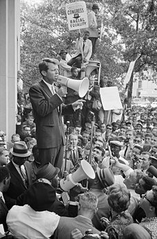 Robert Kennedy, alors procureur général, parlant à une assemblée à l'aide d'un mégaphone devant le siège du département de la Justice des États-Unis, le 14 juin 1963. Une pancarte pour le Congress of Racial Equality est visible. (définition réelle 3 239 × 4 936)