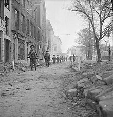 Des hommes avancent avec précaution en ligne, vers la caméra, le long d'une rue en ruines. Ils sont lourdement équipés d'armes et munitions.